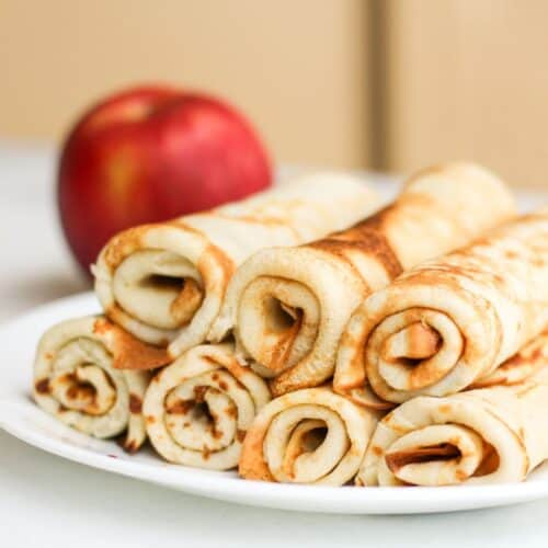 Rolled apple cinnamon crepes on a plate with a fresh red apple blurred in the background.