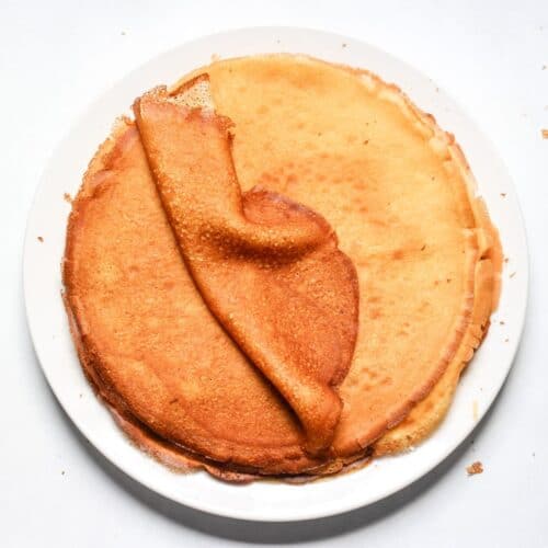 Thin French-style crepes stacked on a plate with the top crepe folded to show the elasticity.
