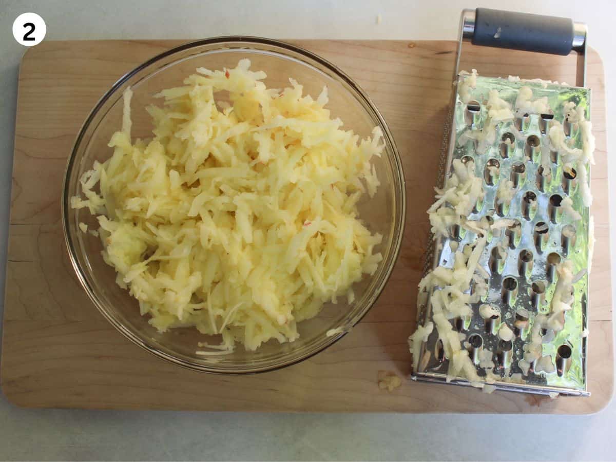 Grated apples in a large glass bowl, box grater next to it. 