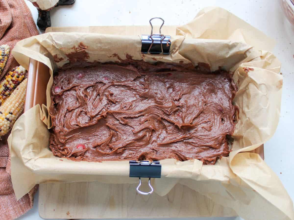 Chocolate batter with folded cranberries evenly spread in a loaf pan lined with parchment paper.