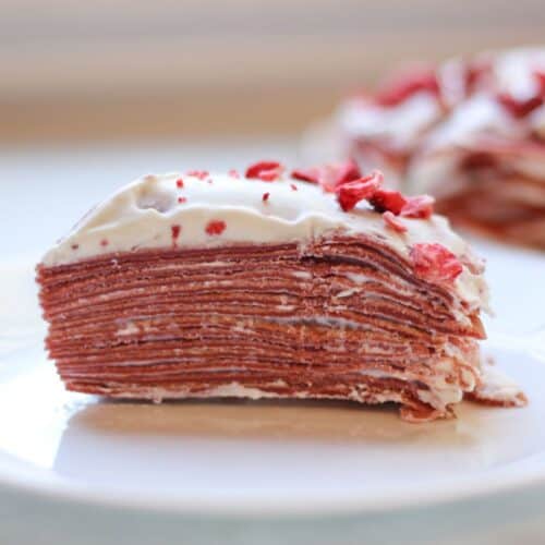 A slice of red velvet crepe cake with creamy frosting on a white plate.