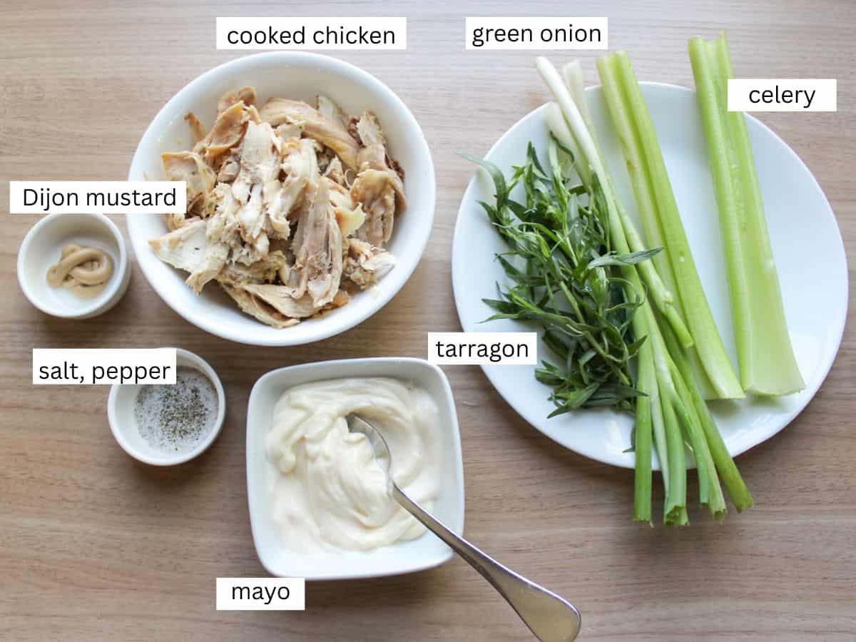 Recipe ingredients on a wooden surface for making classic chicken salad, including cooked chicken, celery, green onions, fresh tarragon, mayo, Dijon mustard, salt and pepper. 