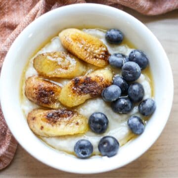 Bowl of creamy cream of buckwheat porridge, topped with caramelized banana sliced, fresh blueberries and drizzle of honey.