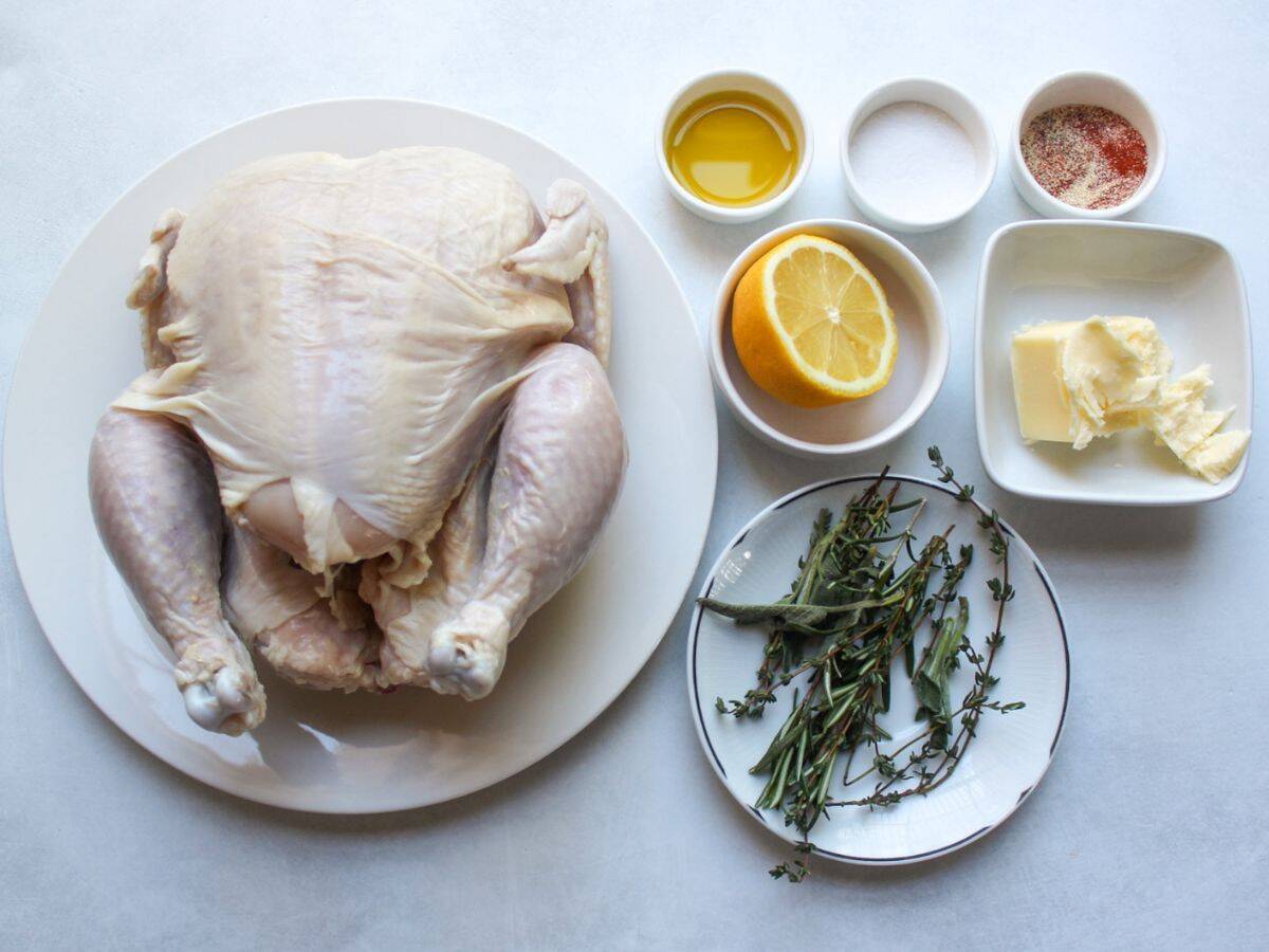 Whole chicken, half of the lemon, fresh herbs, oil, butter, salt and seasonings on a white background.