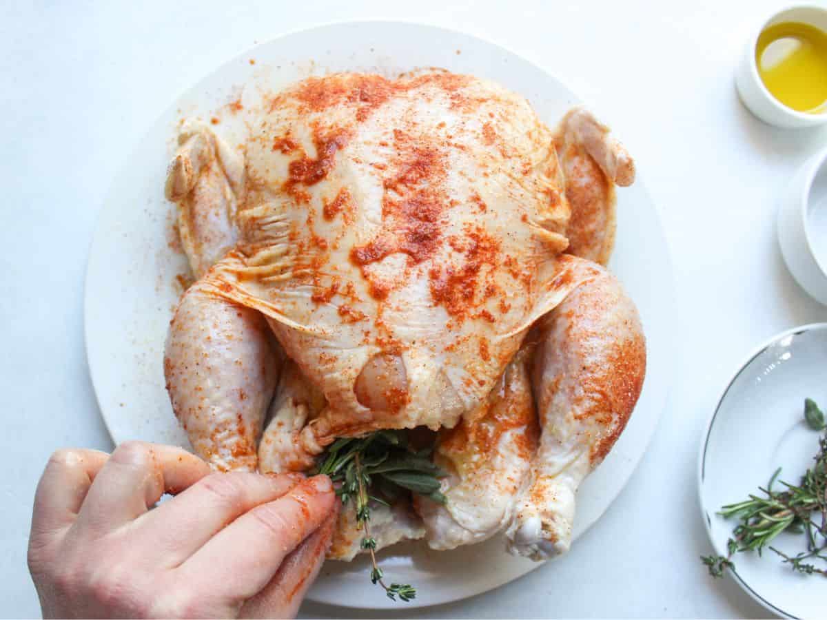 Whole raw chicken coated with olive oil, seasoned with salt, paprika, pepper. Fresh herbs are added to the cavity.