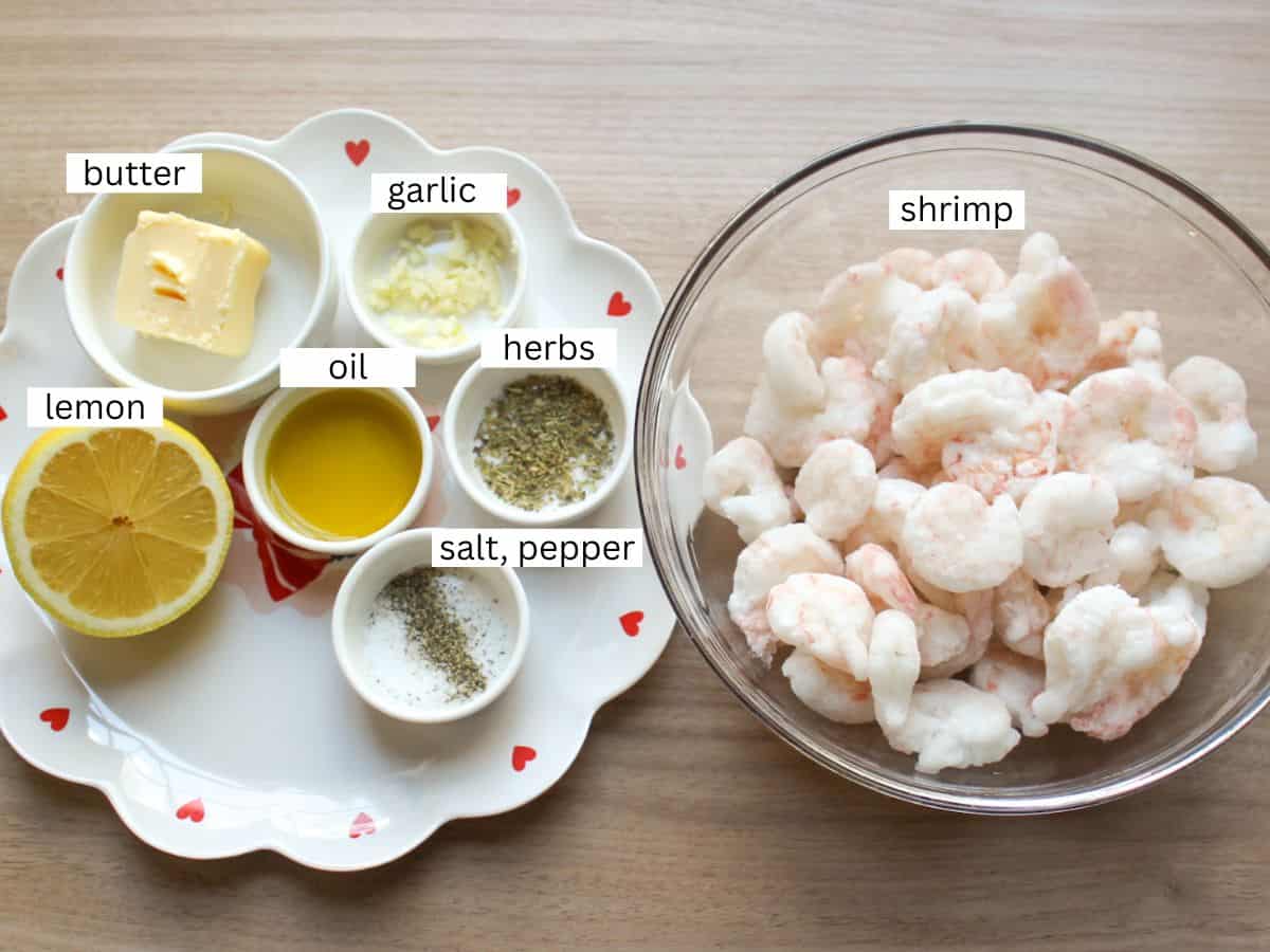 Pan-fried shrimp recipe ingredients including shrimp, oil, butter, lemon, seasonings in separate bowls on a wooden surface. 