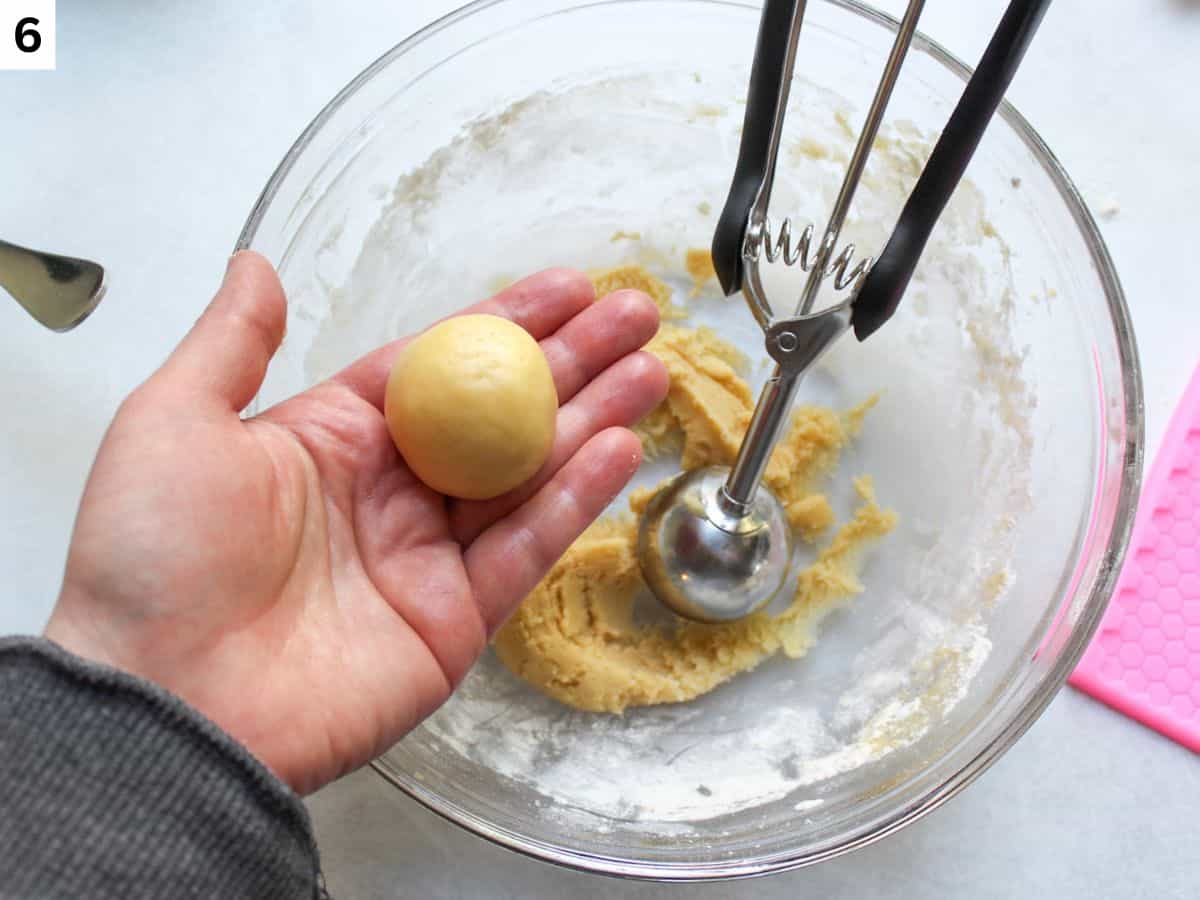 A cookie dough ball  held in hand over a bowl of remaining dough. 