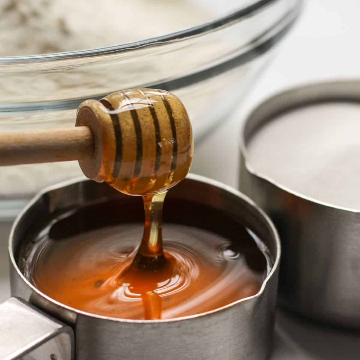 Measuring cup filled with honey with a wooden honey dipper next to bowl flour and sugar.
