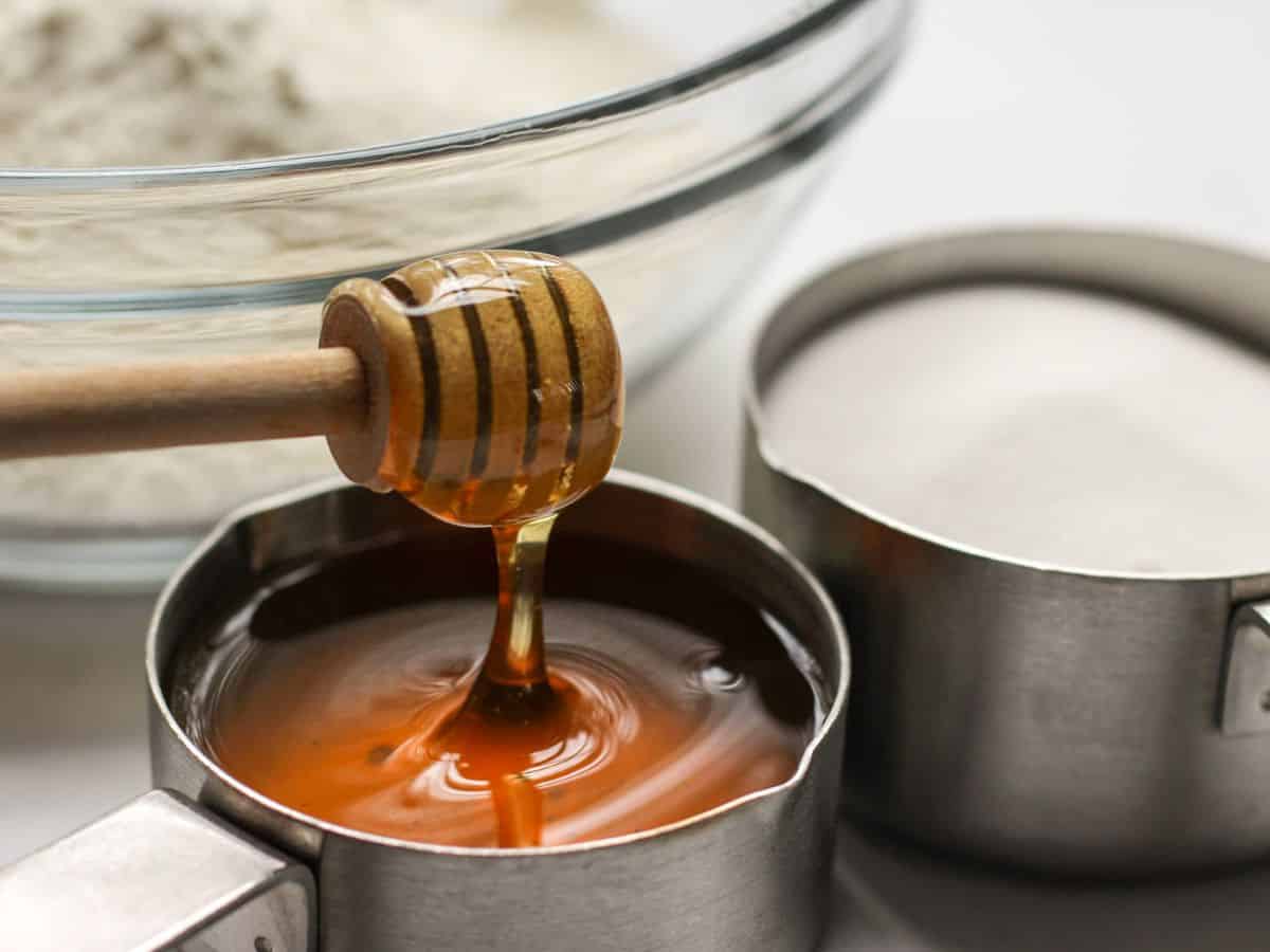 Measuring cup filled with honey with a wooden honey dipper next to bowl flour and sugar. 