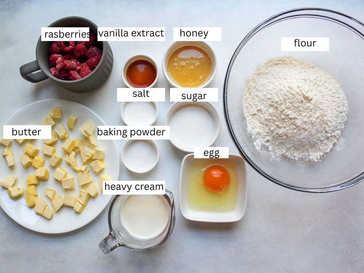 Ingredients for honey raspberry scones, including flour, cold cubed butter, heavy cream, a bowl of honey and frozen raspberries. 