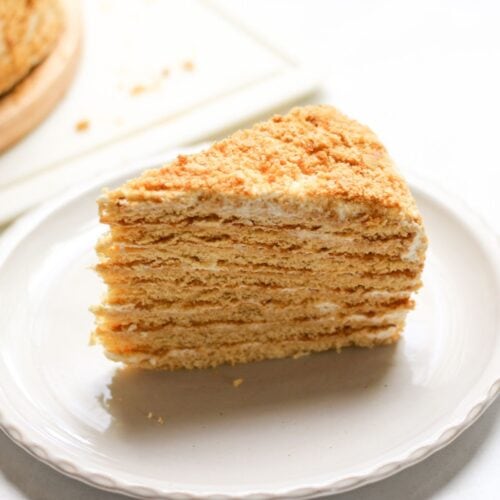 Authentic 8-layer Medovik honey cake on a white plate, showing the soft hydrated layers, sour cream frosting, and a signature golden crumb coating.