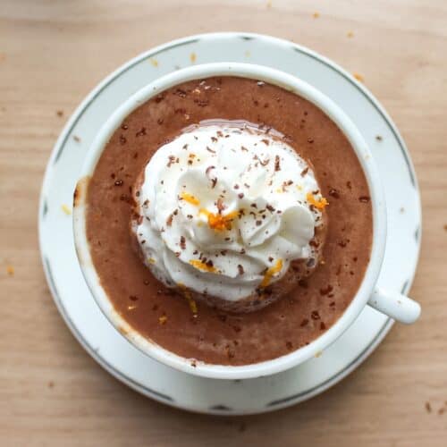 A mug of thick, rich European hot chocolate topped with a swirl of whipped cream, an orange peel garnish, and grated dark chocolate.
