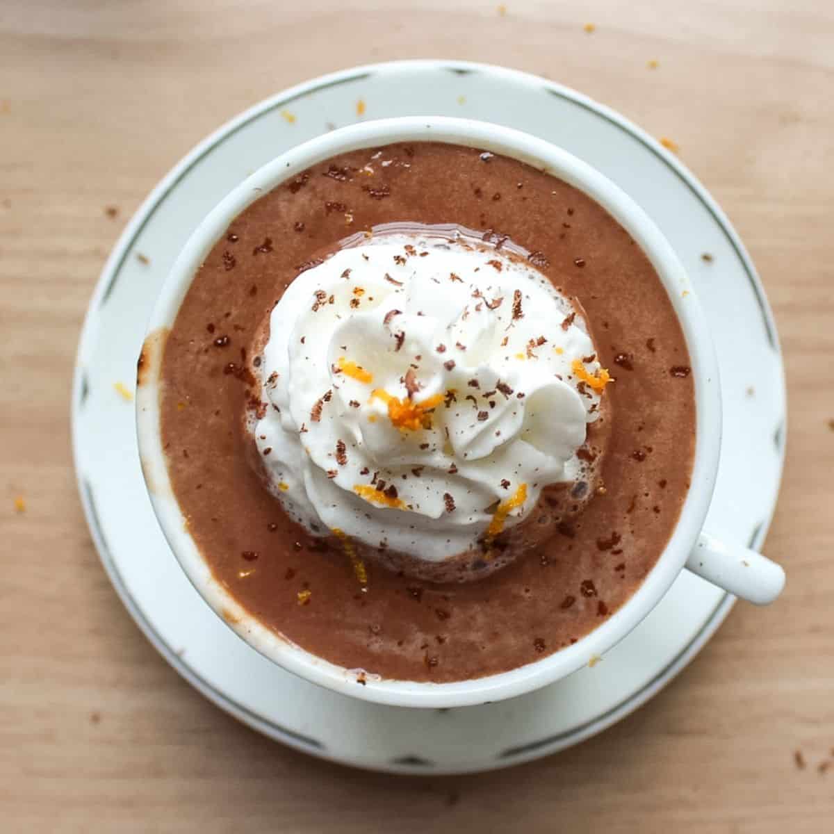 A mug of thick, rich European hot chocolate topped with a swirl of whipped cream, an orange peel garnish, and grated dark chocolate.