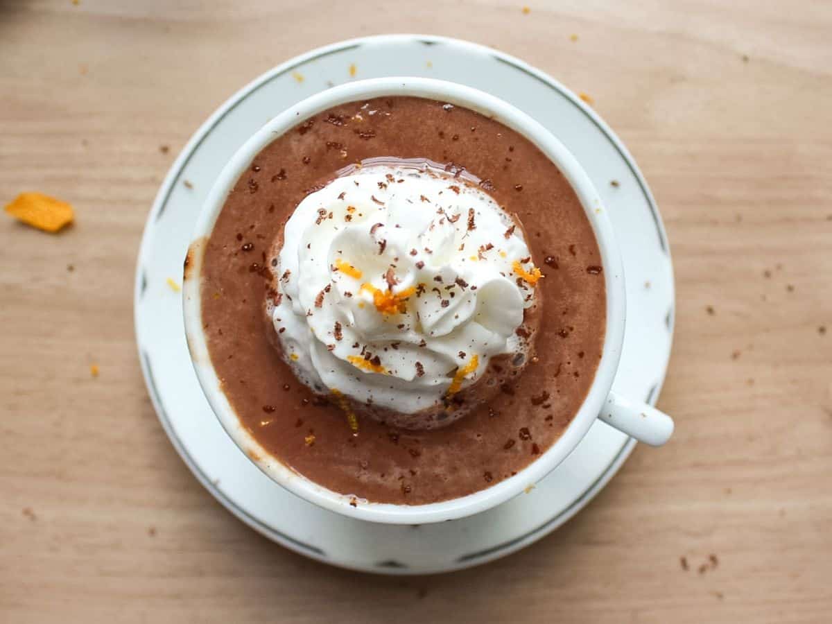 A mug of thick, rich European hot chocolate topped with a swirl of whipped cream, an orange peel garnish, and grated dark chocolate.