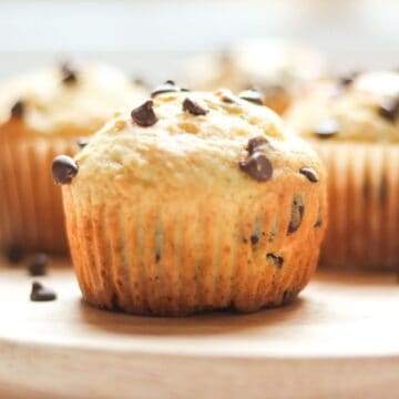 A freshly baked sour cream chocolate chip muffin removed from its paper liner, showing a tall domed top and golden-brown edges on a wooden tray.
