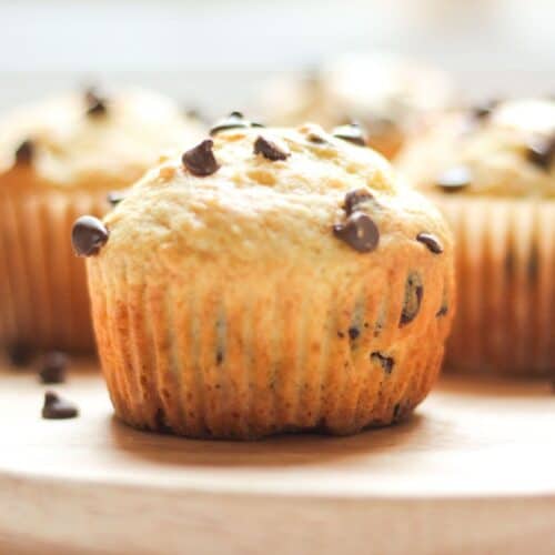 A freshly baked sour cream chocolate chip muffin removed from its paper liner, showing a tall domed top and golden-brown edges on a wooden tray.