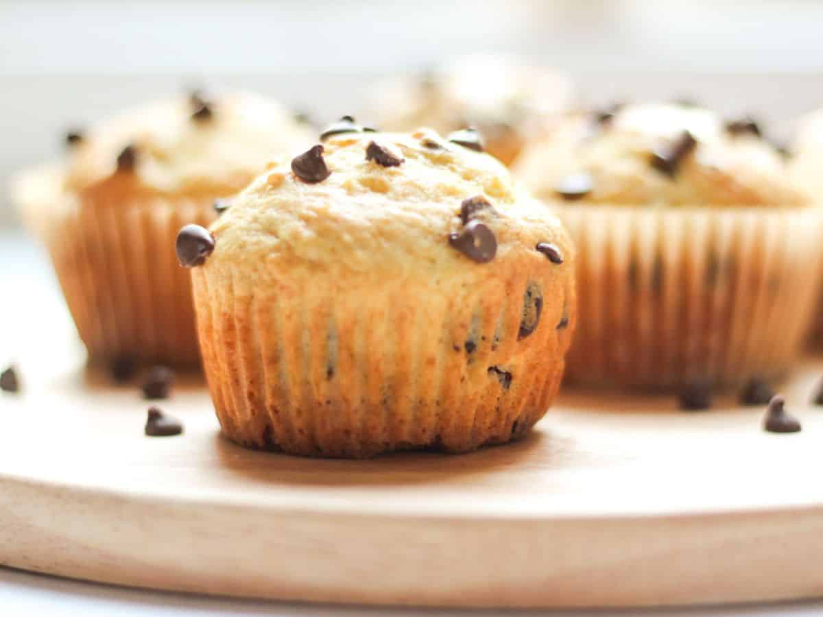 A close-up of a golden, unwrapped sour cream chocolate chip muffin sitting on a wooden board, surrounded by scattered chocolate chips with more muffins in the background.