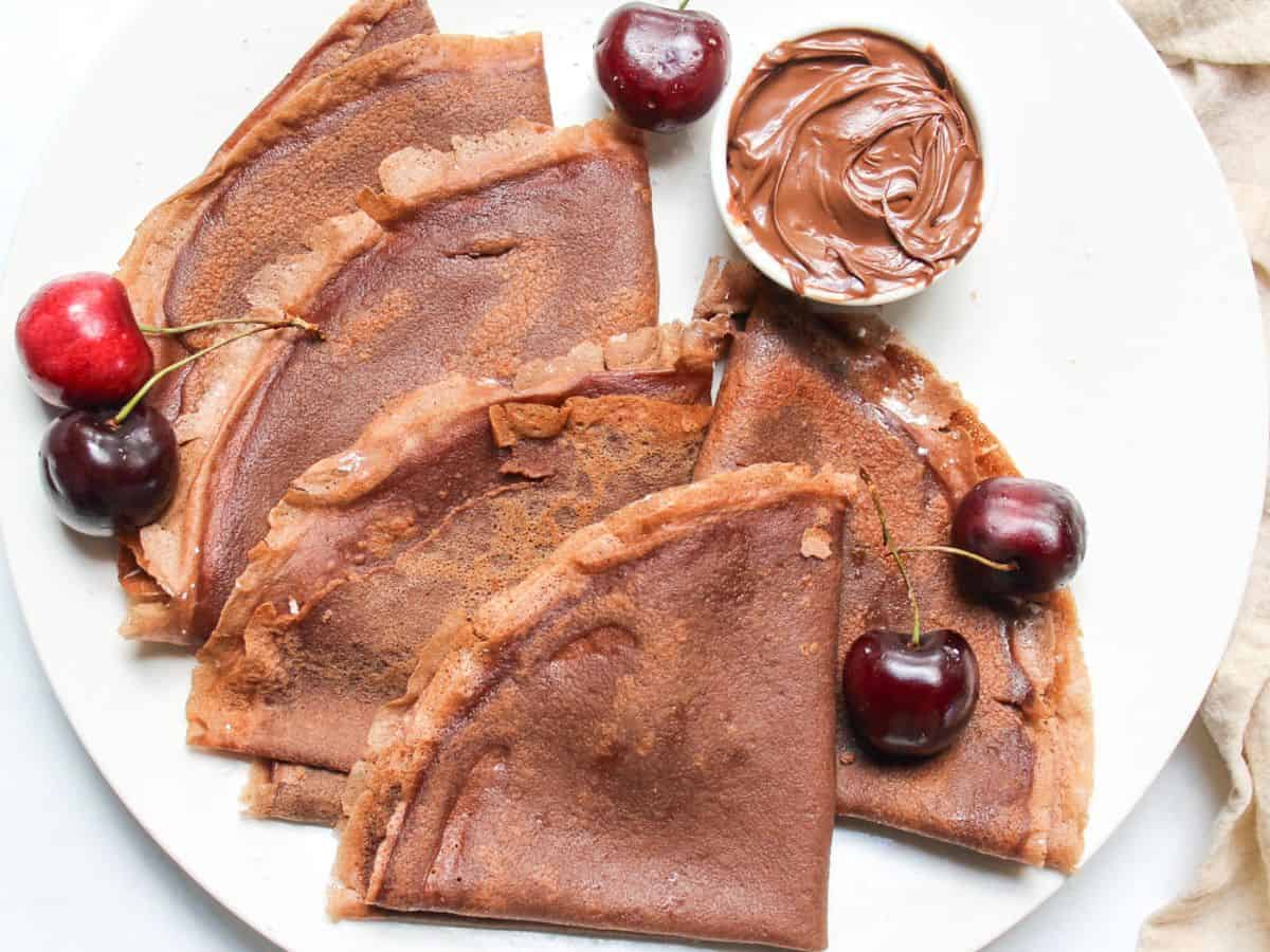 Folded chocolate crepes served on a white plate with fresh sweet cherries and a small ramekin with chocolate hazelnut spread.