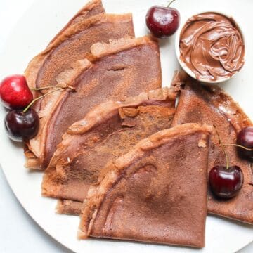 Folded chocolate crepes served on a white plate with fresh sweet cherries and a small ramekin with chocolate hazelnut spread.
