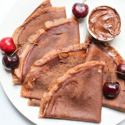Folded chocolate crepes served on a white plate with fresh sweet cherries and a small ramekin with chocolate hazelnut spread.