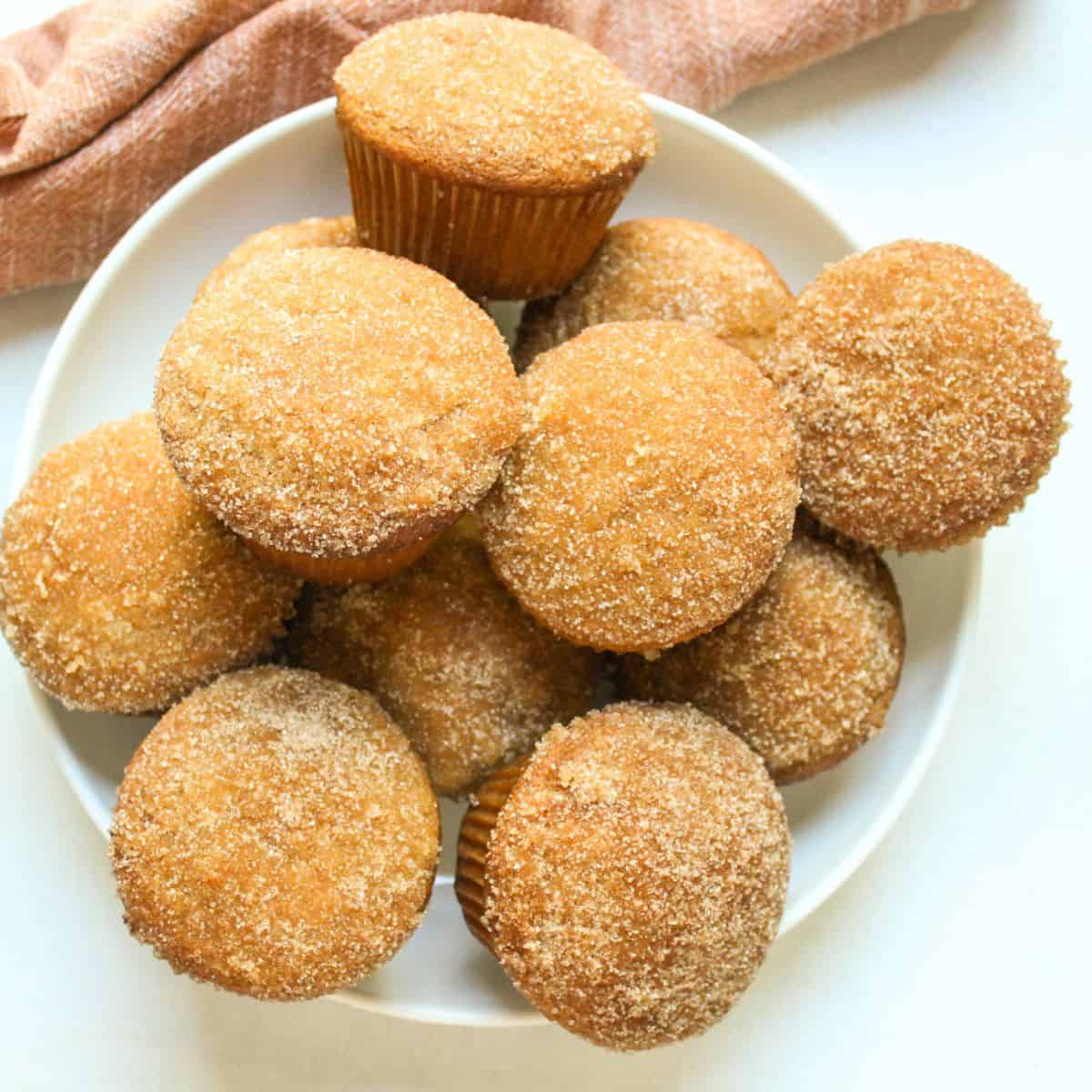 Tall homemade apple cider muffins with sparkling cinnamon-sugar donut coatings piled high on a white serving plate.