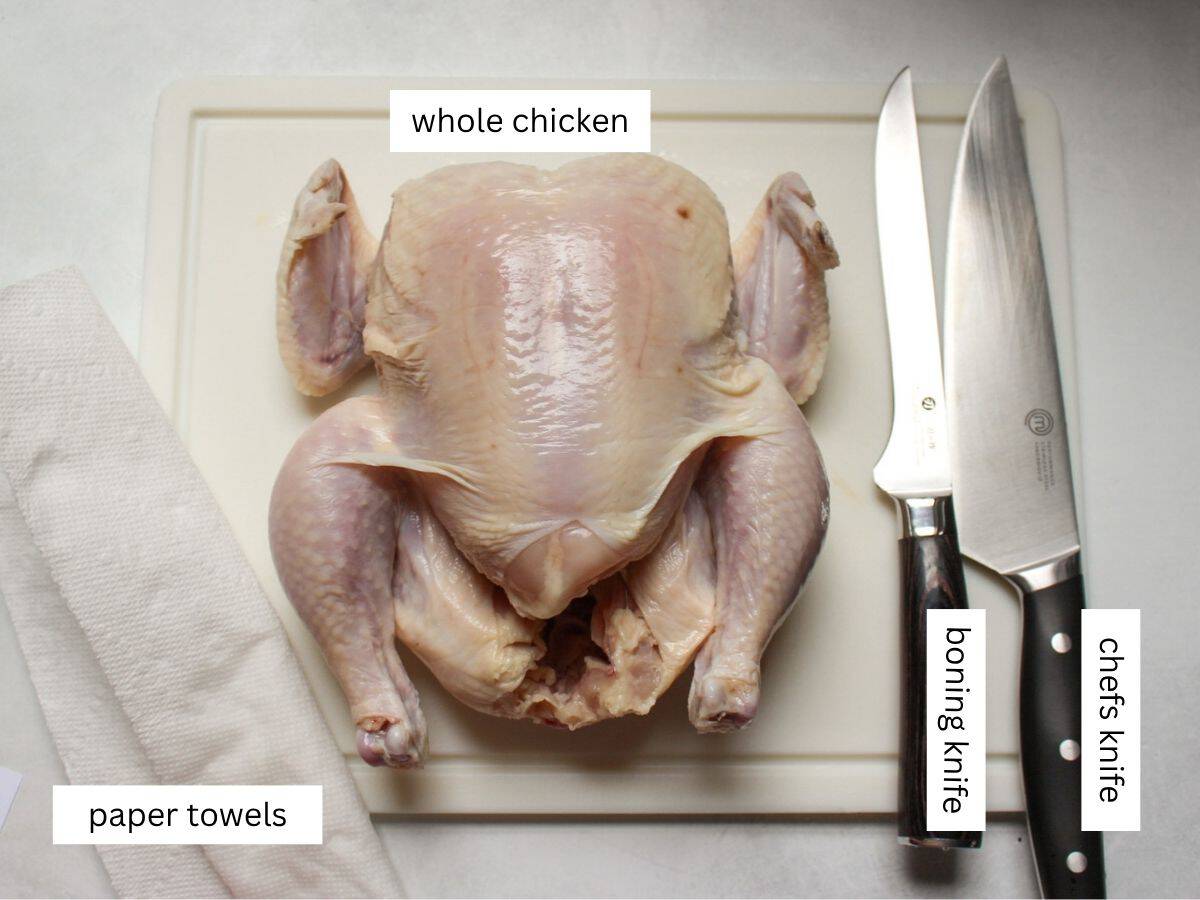 Whole chicken, boing knife, chefs knife and paper towels on a white plastic cutting board.